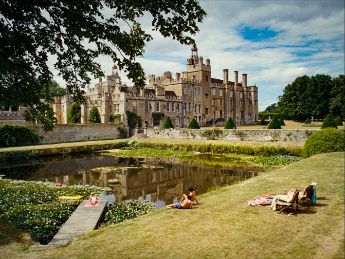 A screenshot from Saltburn. In a wide angle shot, you see the entirety of the Salturn estate and the part of the grounds that overlook an artificial lake. Three figures rest by the lakeside.
