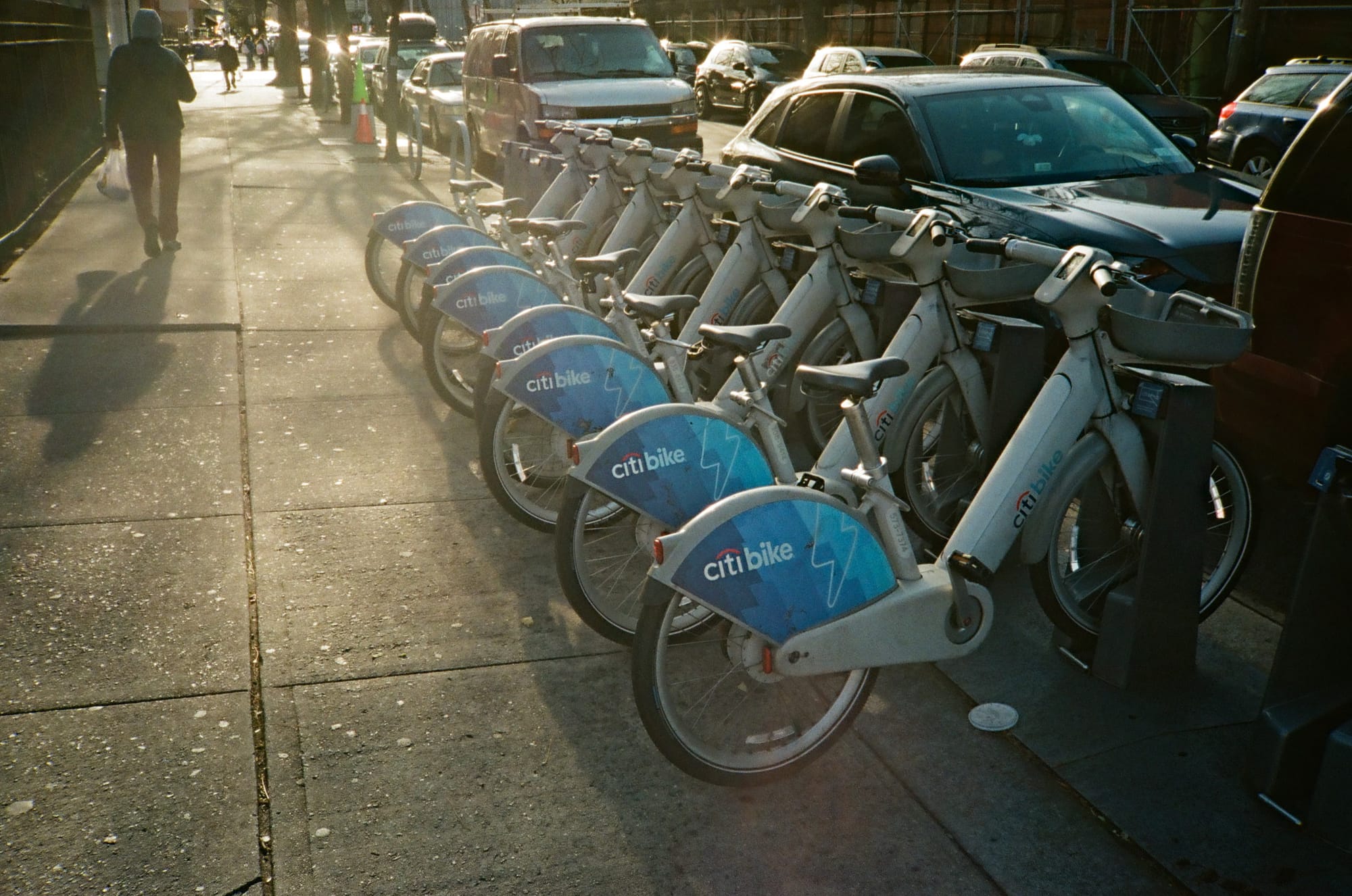 A rack of Citi Bikes at sunset, the haze on a film photo of the rack as a person in the back walks away.