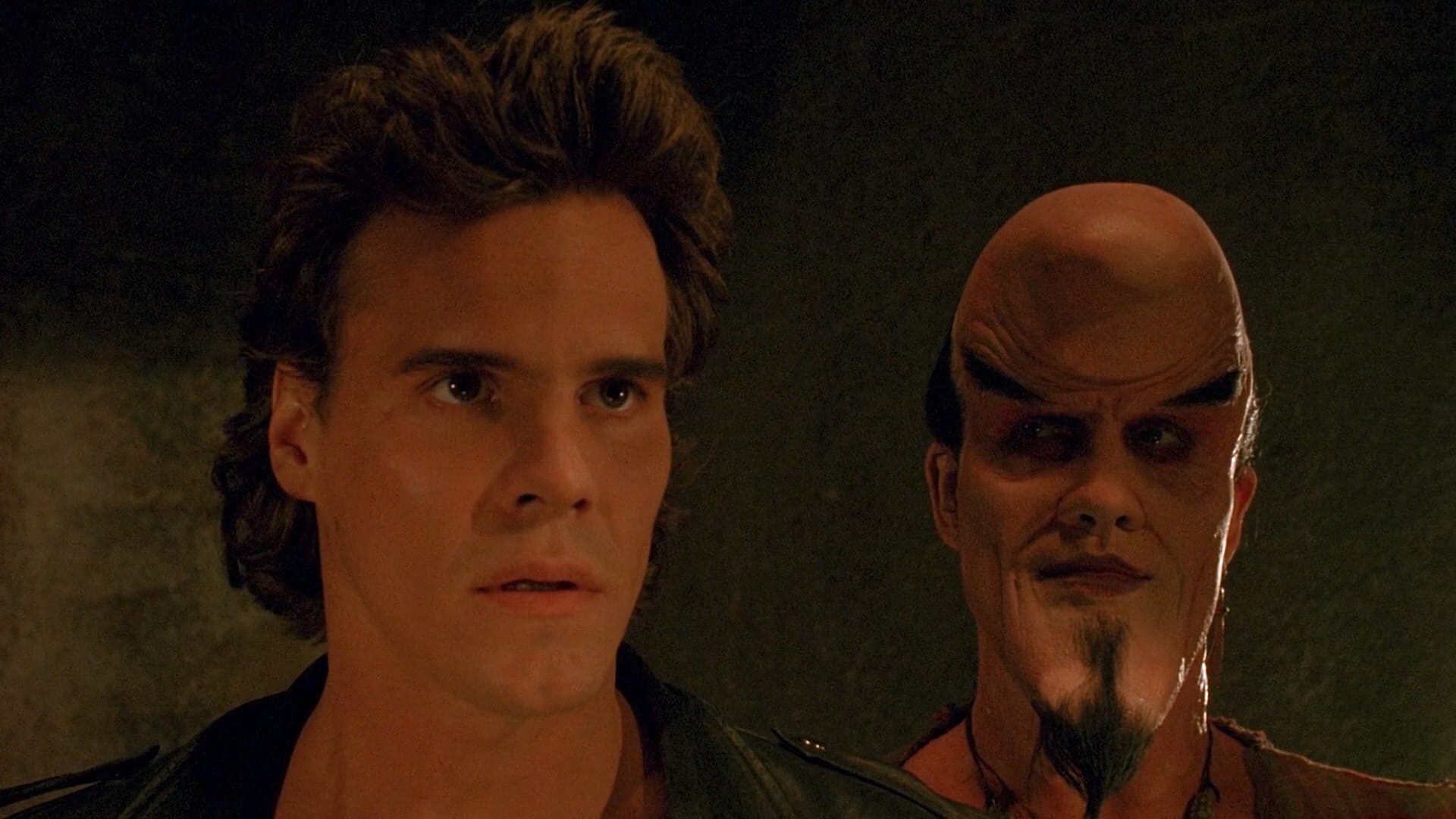 A normal looking man in a leather jacket, standing next to a man whose face is distorted into the shape of a moon, with strong eyebrows and a devil goatee.